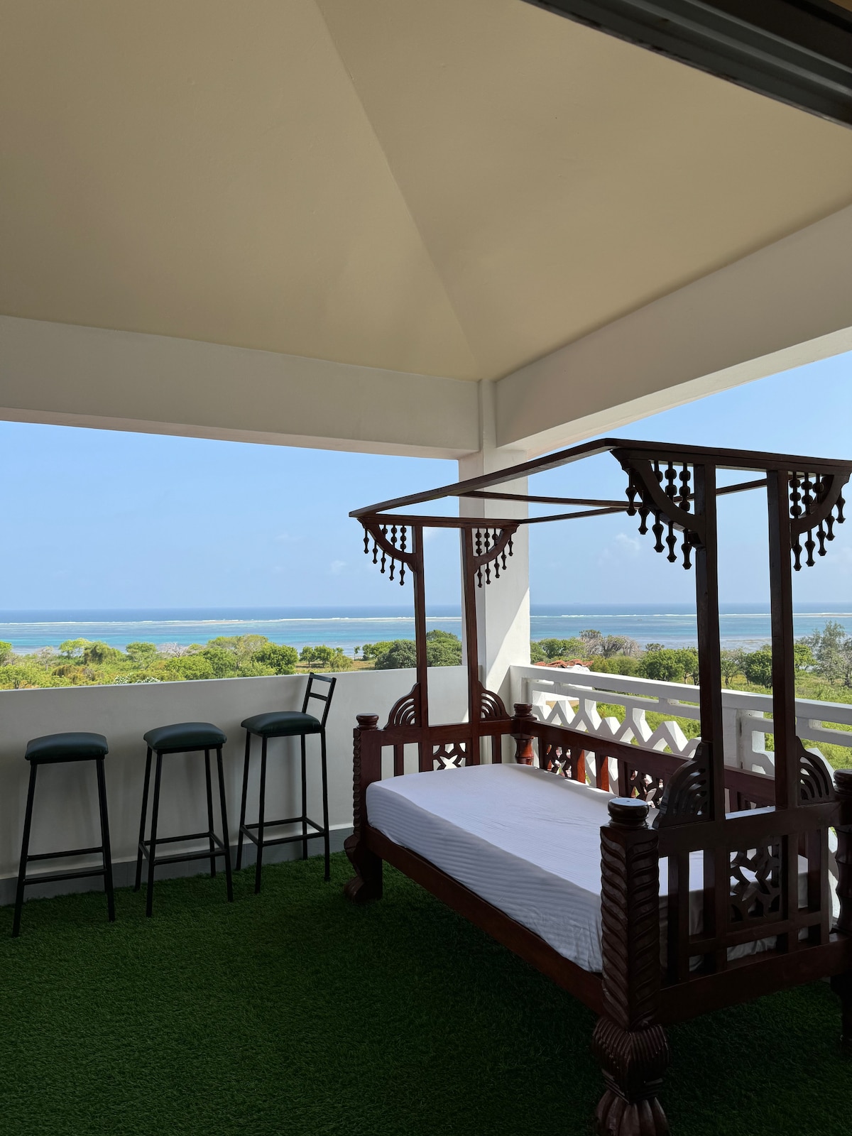 A private balcony is shown with a traditional wooden daybed, positioned for ocean views. Two high chairs are placed along the edge. Lush green grass flooring enhances the serene environment, while the horizon displays a clear blue sky meeting the water.