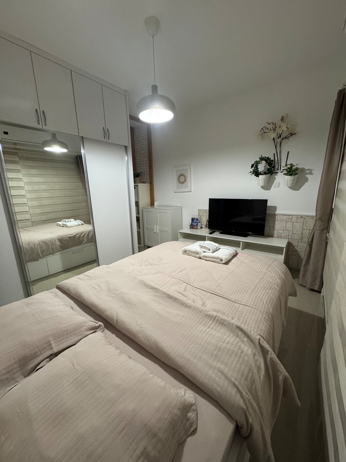 A cozy bedroom is presented with a spacious bed covered in soft linens. A flat-screen television rests on a light-colored stand, while a mirrored wardrobe provides storage. Natural light enters through a window dressed in subtle curtains, and decorative plants add a touch of greenery.