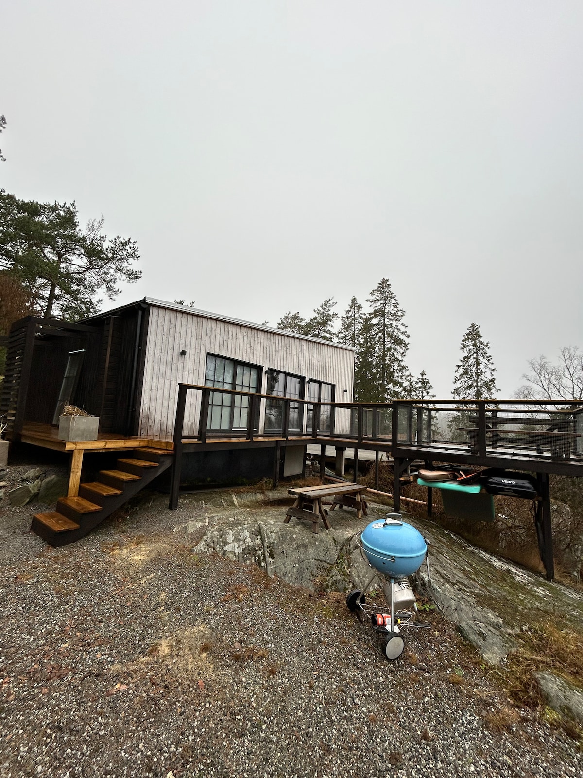 A modern retreat is set on a rocky hillside, featuring a wooden deck and steps leading to the entrance. The exterior showcases a combination of sleek materials and large windows, surrounded by trees, providing a natural, tranquil setting.