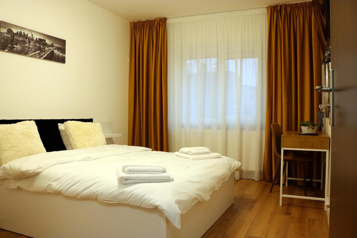 A minimalist bedroom features a spacious bed adorned with neatly arranged towels. Natural light filters through sheer and dark curtains, casting a soft glow. A simple desk and chair are positioned nearby, complementing the warm wooden flooring.