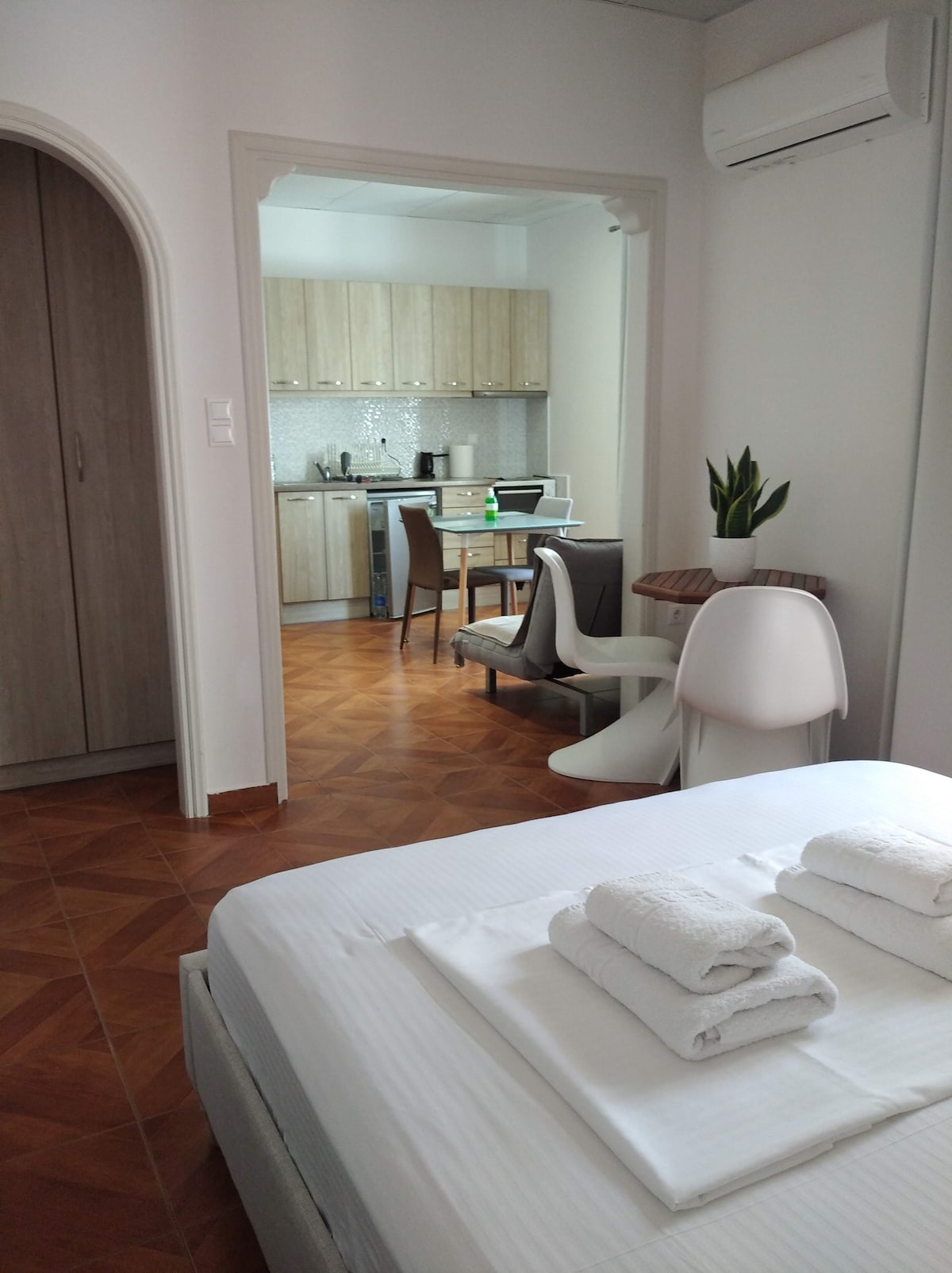 A well-lit interior captures a cozy sleeping area featuring a spacious bed with neatly arranged towels. In the background, a modern kitchenette is visible, accompanied by a dining table and chairs, enhancing the functionality of the space. The warm wood tones of the flooring add to the inviting atmosphere.