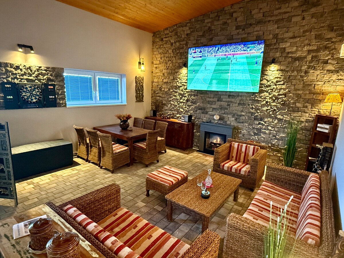 A spacious living area features a stone wall accent and a large television mounted above a fireplace. Woven furniture includes a cozy seating arrangement with striped cushions and a wooden dining table accommodating multiple seats. Natural light is provided by nearby windows.