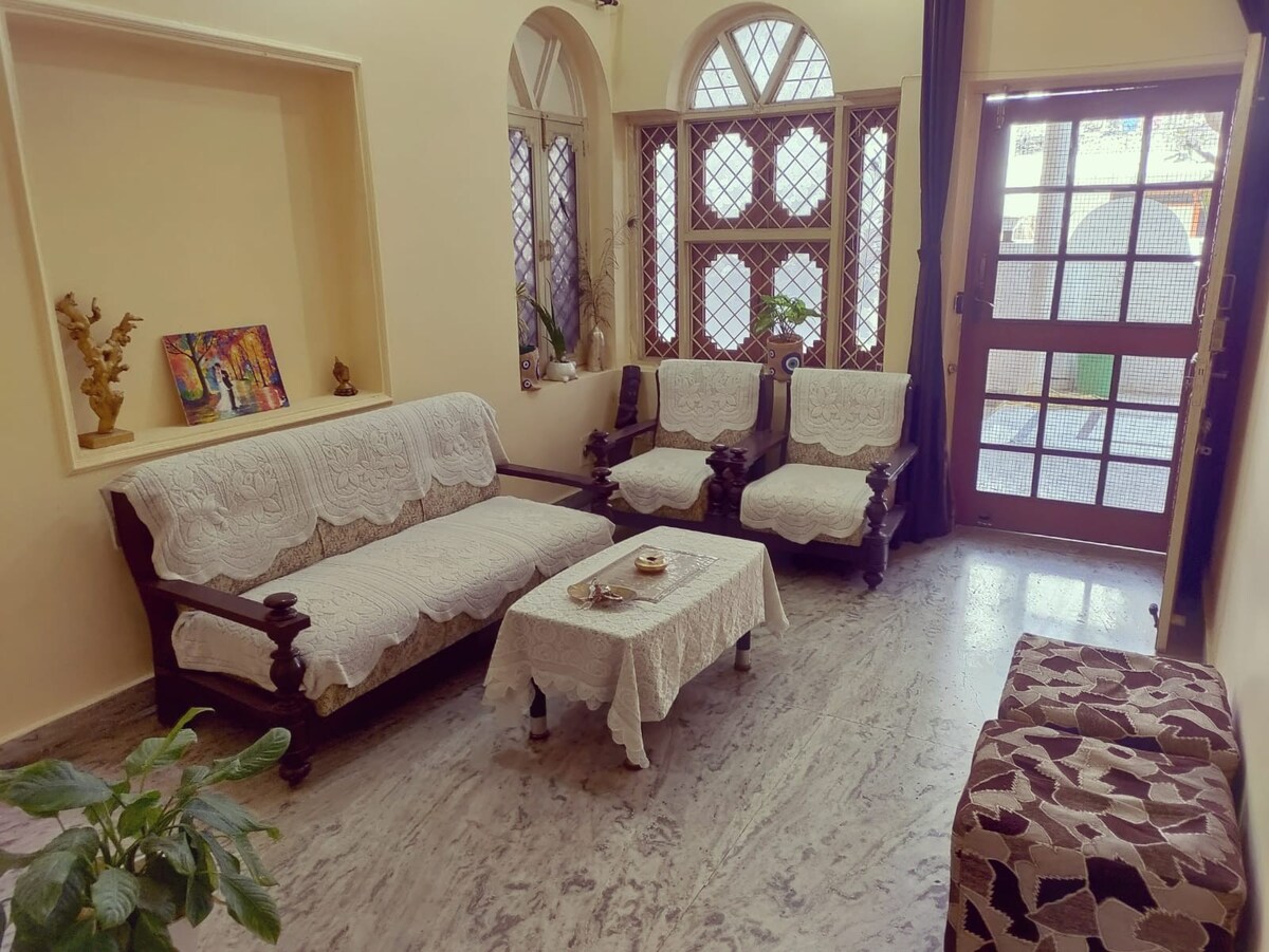 A drawing room features a set of elegant seating arrangements, including two armchairs and a sofa, all draped with light-colored covers. A small coffee table is centered in front of the seating area. Natural light filters through the large windows, creating a welcoming environment.