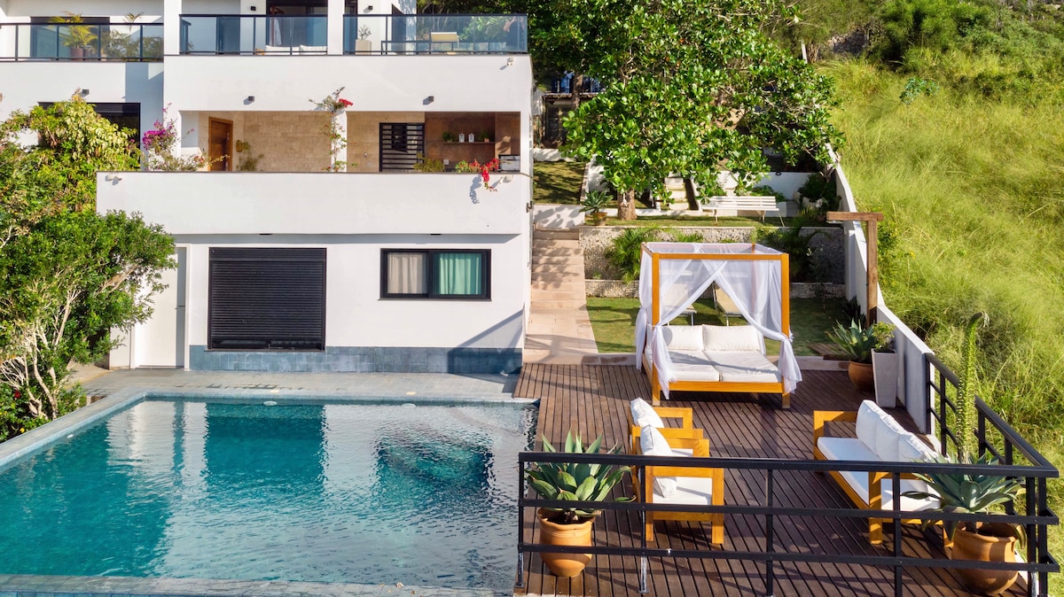 An outdoor area features a shared infinity pool surrounded by greenery. A shaded seating area with lounge chairs is present, alongside natural wooden elements. The space is designed for relaxation, with easy access to steps leading to the house and its scenic environment.