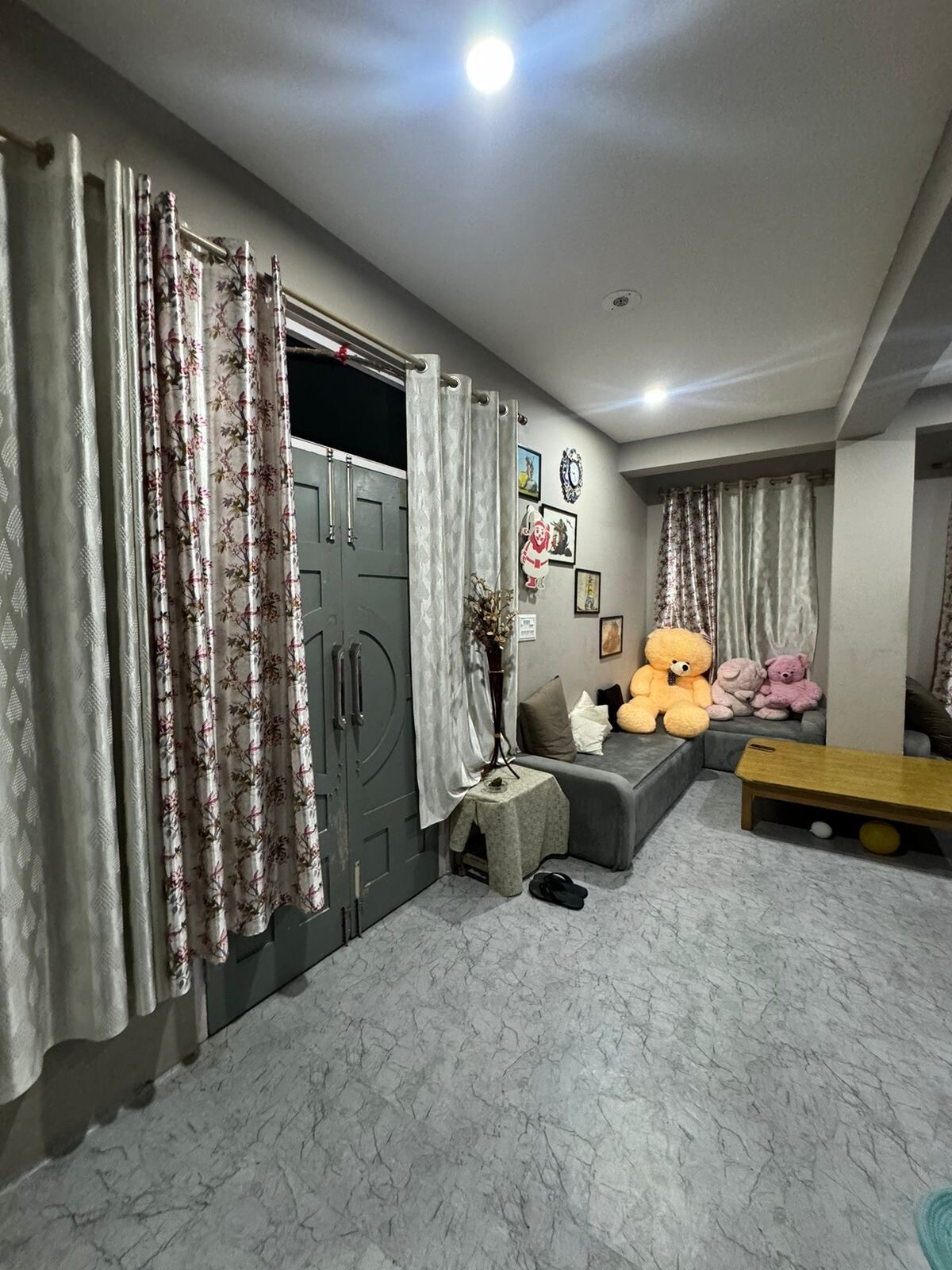 A living area features soft lighting and a neutral color palette. A large sofa is accompanied by decorative plush toys, while a wooden coffee table occupies the center space. Floral curtains frame the windows, adding a touch of elegance to the room.