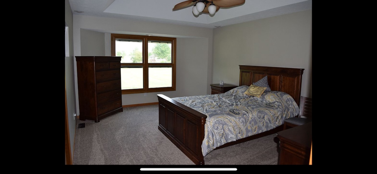 A spacious bedroom features a large wooden bed with a patterned quilt and coordinating pillows. Light enters through a wide window with a view of the greenery outside. A tall dresser and a nightstand are positioned nearby, complimenting the room's neutral tones.