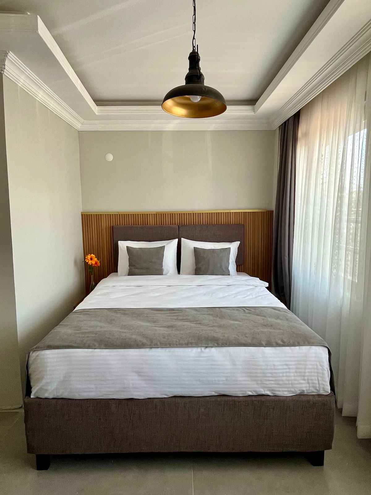 A cozy bedroom features a spacious bed dressed in white linens with grey accents. Natural light filters through sheer curtains, illuminating the room. A decorative flower adds a touch of color, while a minimalist pendant light hangs from the ceiling, enhancing the modern design.