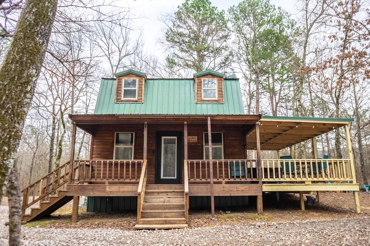 A rustic cabin with a green metal roof is surrounded by trees. The entrance features a set of wooden steps leading to a covered porch, which extends to one side. Two small windows are visible on the upper level, enhancing the cabin's inviting facade.