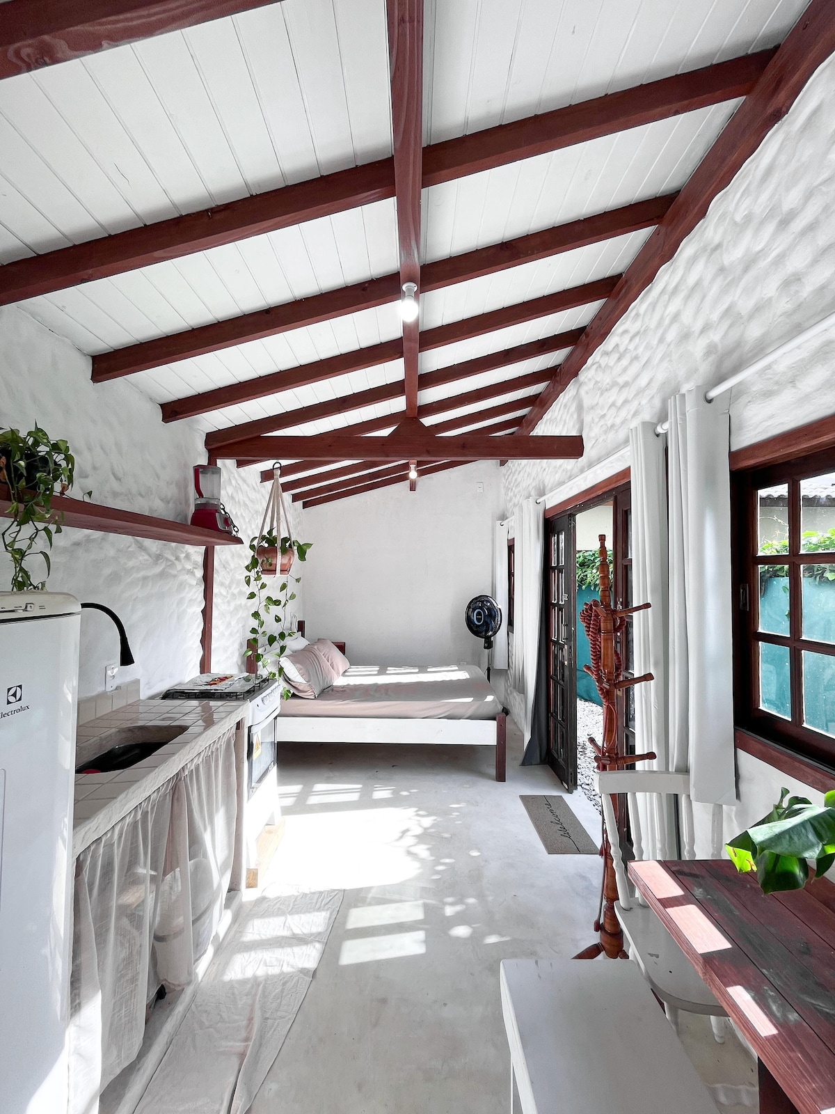 An open-concept space is shown with a cozy sleeping area beside a kitchen. Natural light floods in through windows, highlighting the textured white walls and wooden beams. Indoor plants add a touch of greenery, creating a calming atmosphere.