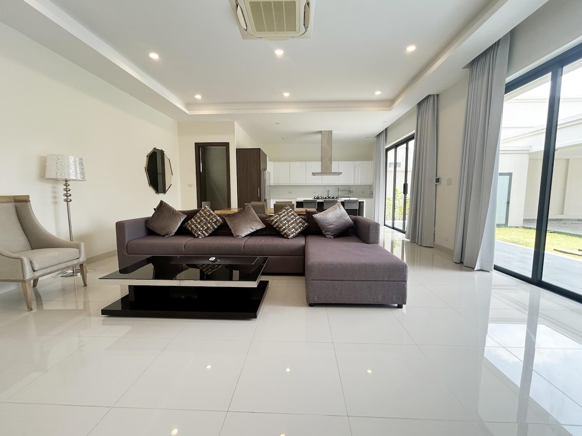 A spacious living area is equipped with a large sectional sofa adorned with decorative cushions. A sleek coffee table sits in front, complementing the modern aesthetic. Large glass doors provide natural light and views of the outdoor space, while neutral-toned walls enhance the open feel.