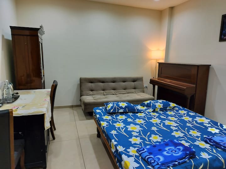 Ოთახი ლურჯი ყვავილების თეთრეულით დაფარული საწოლით, რბილი ნაცრისფერი ფუტონი, ხის თავდახურული ფორტეპიანო, კუთხეში იატაკის მაღალი ლამპა, სასადილო მაგიდა სკამებით და გარდერობი შორეულ კედელთან.