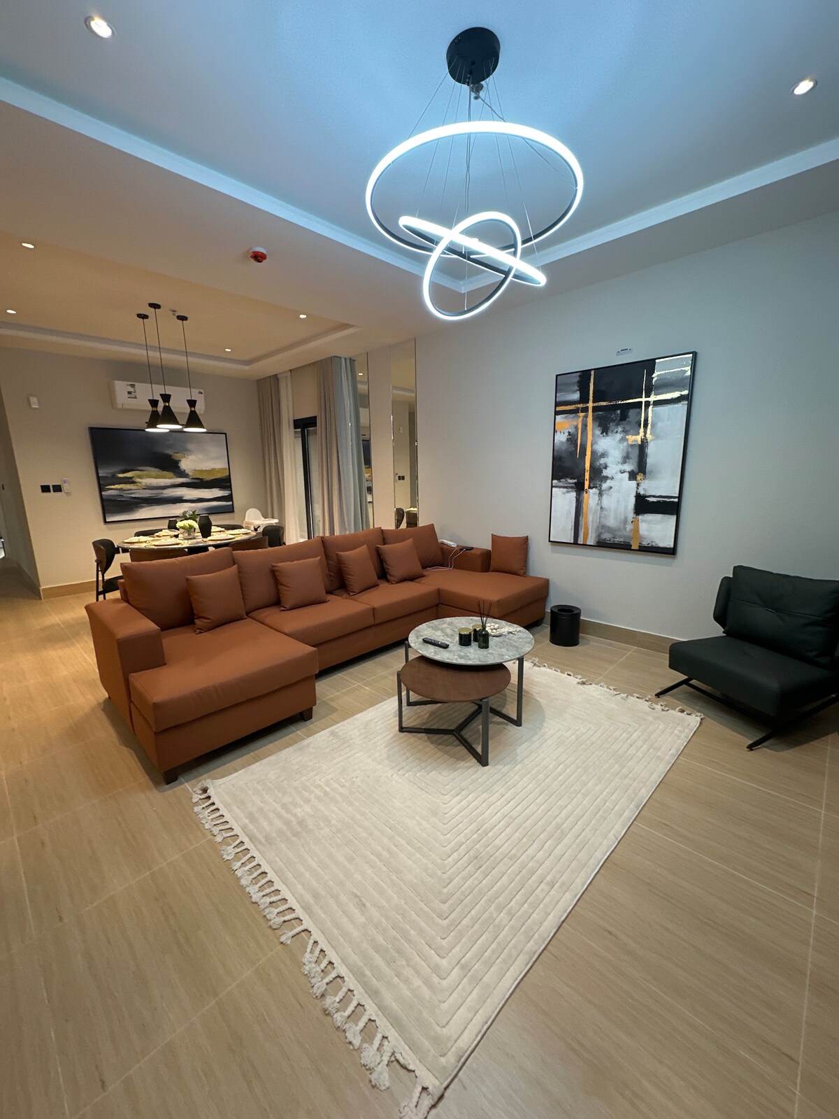 The living room features a spacious sectional sofa in warm tones, complemented by a round table at its center. A textured area rug lies underneath, and a sleek black chair is positioned nearby. Modern lighting fixtures illuminate the space, enhancing the overall ambiance.