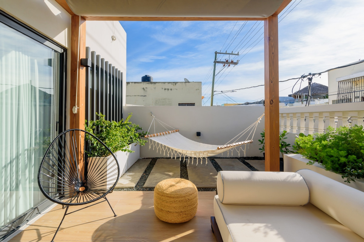 A relaxing terrace is highlighted by a swinging hammock, surrounded by greenery. An inviting lounge chair and a circular pouf complement the space, which is framed by wooden beams. Light flows in through large sliding doors, enhancing the connection between indoors and outdoors.
