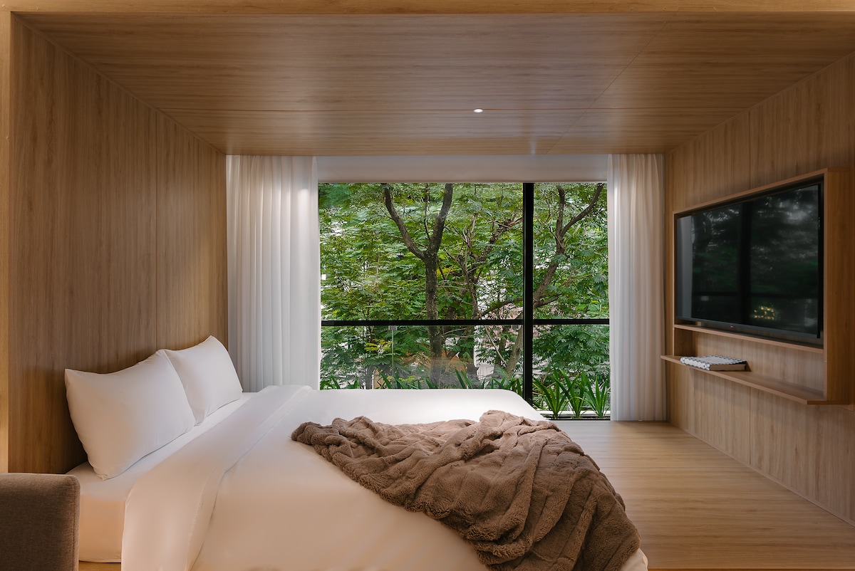 The sleeping area features warm wooden paneling and a spacious bed, dressed in white linens with a soft brown throw. Floor-to-ceiling windows frame a view of lush greenery, allowing natural light to fill the room.