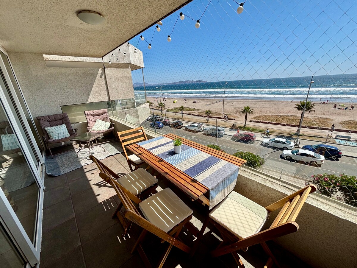 A well-furnished balcony with a coastal view features a wooden dining table surrounded by folding chairs. Plush lounge chairs are positioned nearby, with sunlight illuminating the area. A glimpse of the ocean and beach is visible through the glass railing, enhancing the serene atmosphere.