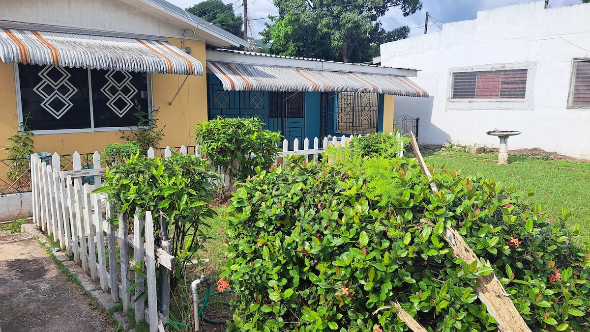 A yellow single-story house is surrounded by lush greenery and a white picket fence. The front yard features well-maintained bushes with blooming flowers, and a stone birdbath is situated in the garden space. Large windows with decorative awnings are present, allowing natural light inside.