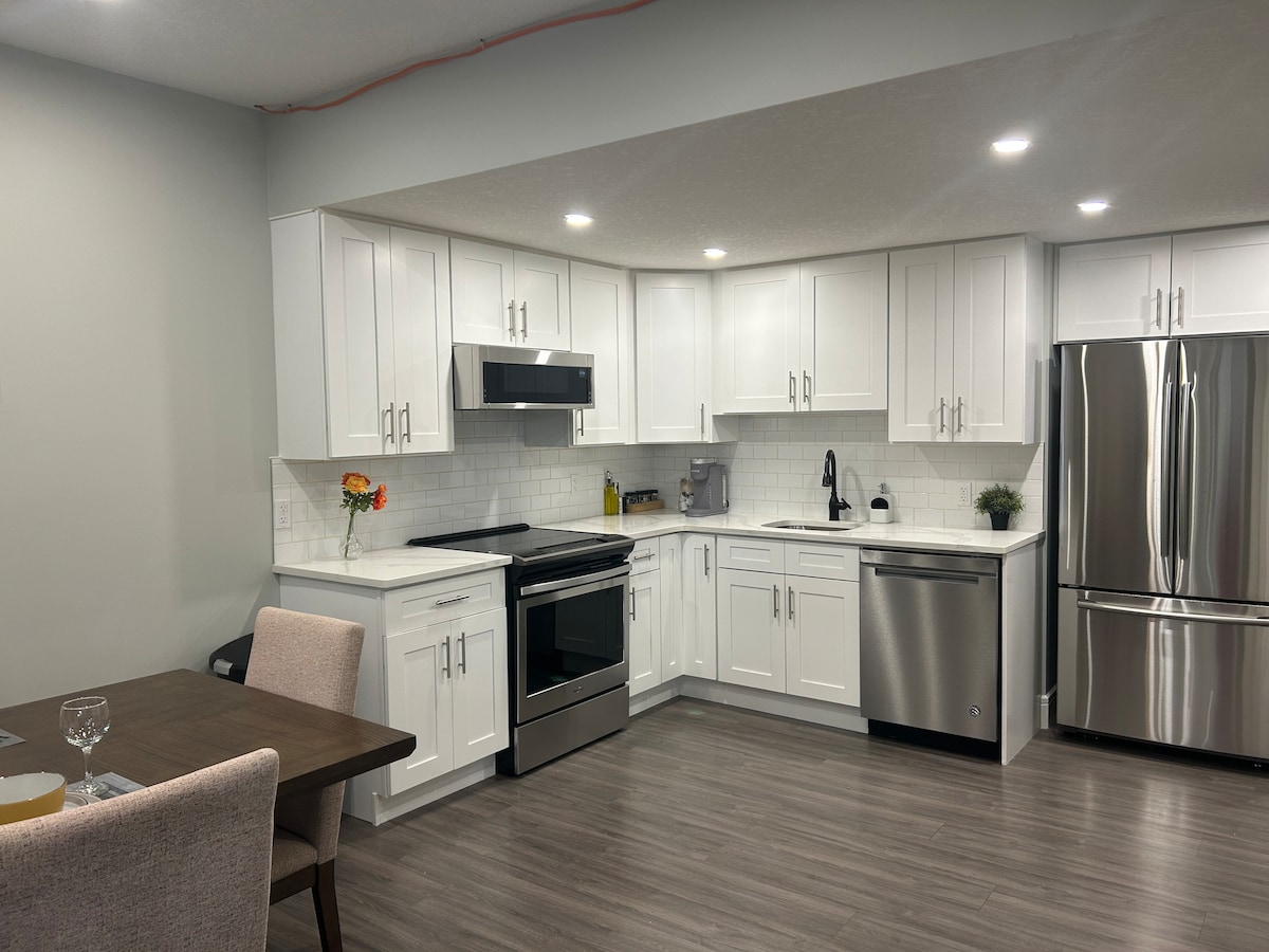 A modern kitchen is featured, equipped with white cabinetry, stainless steel appliances, and a large refrigerator. The space includes a sleek countertop and a sink, while a well-organized dining area with a table and chairs is visible in the foreground.