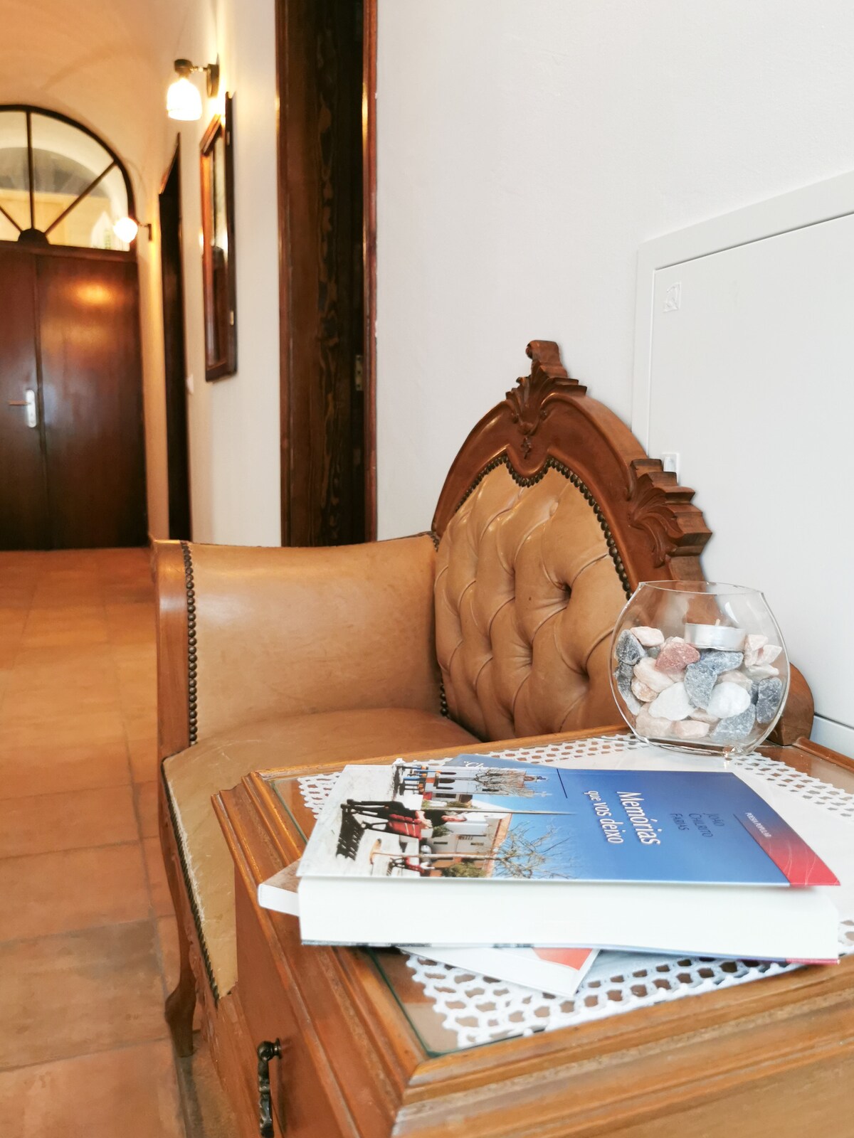 An elegant seating area is highlighted by a vintage armchair with tufted detailing. A wooden side table holds a travel guide and a decorative glass container filled with stones. The hallway, featuring warm lighting and wooden accents, extends in the background.