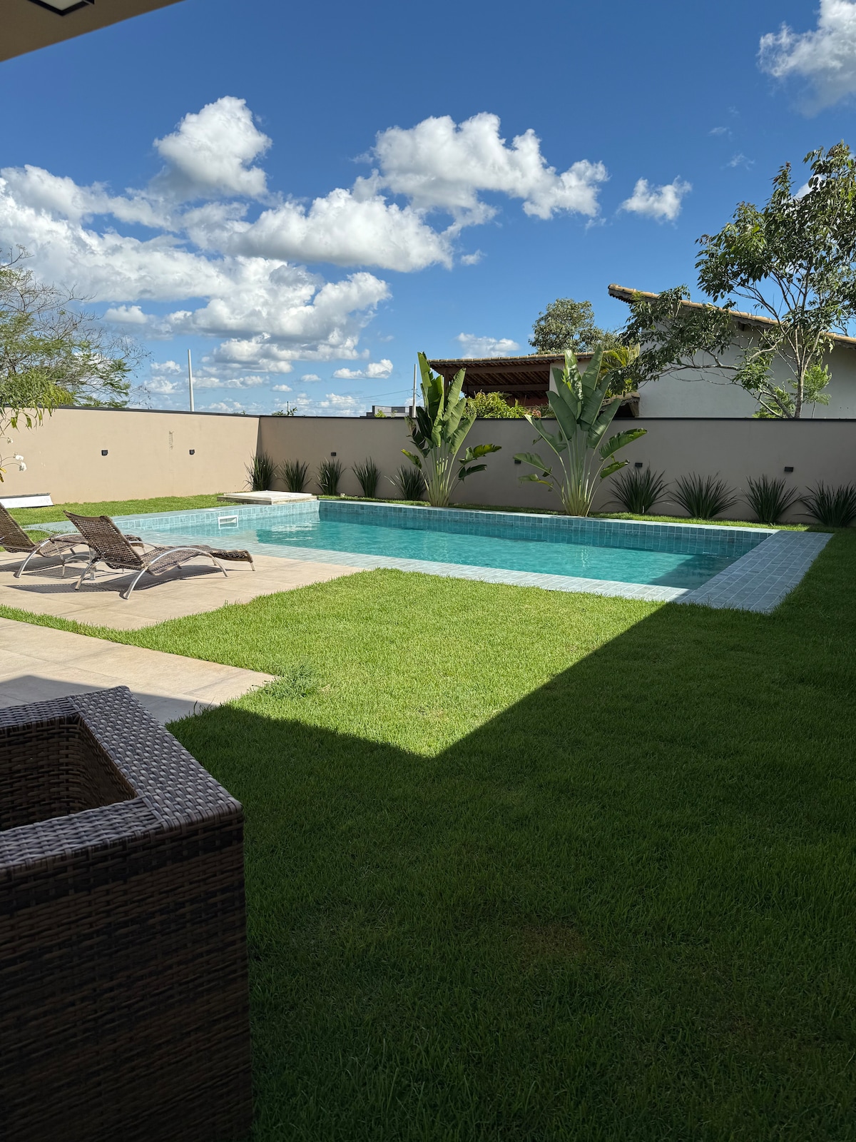 A glistening pool is set amidst a manicured lawn, surrounded by greenery and a wall. Two lounge chairs are positioned near the water's edge, inviting relaxation under the sun. Bright blue skies and scattered clouds create a serene backdrop.