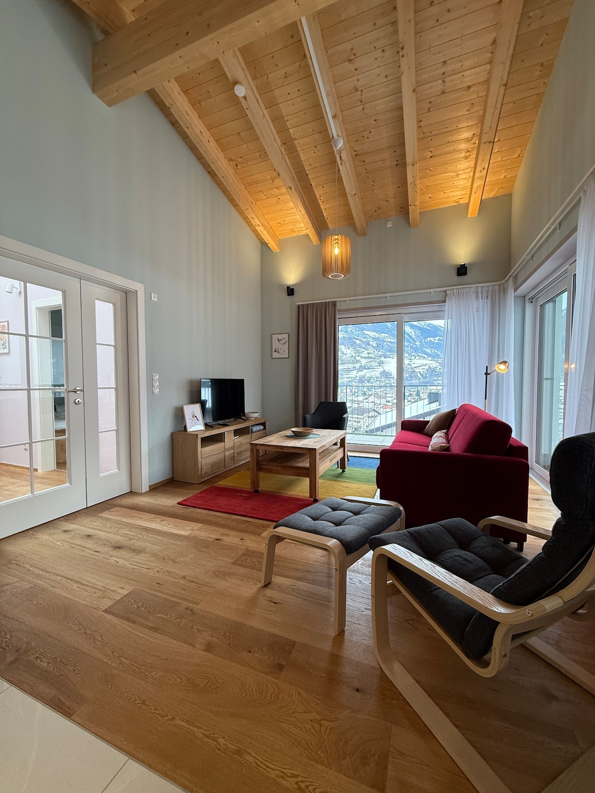 A bright living area features a high, wooden beam ceiling and large glass doors that offer expansive views of the surrounding mountains. Comfortable seating includes a red sofa, a cozy armchair, and a wooden coffee table, complemented by a colorful area rug on warm wood flooring.
