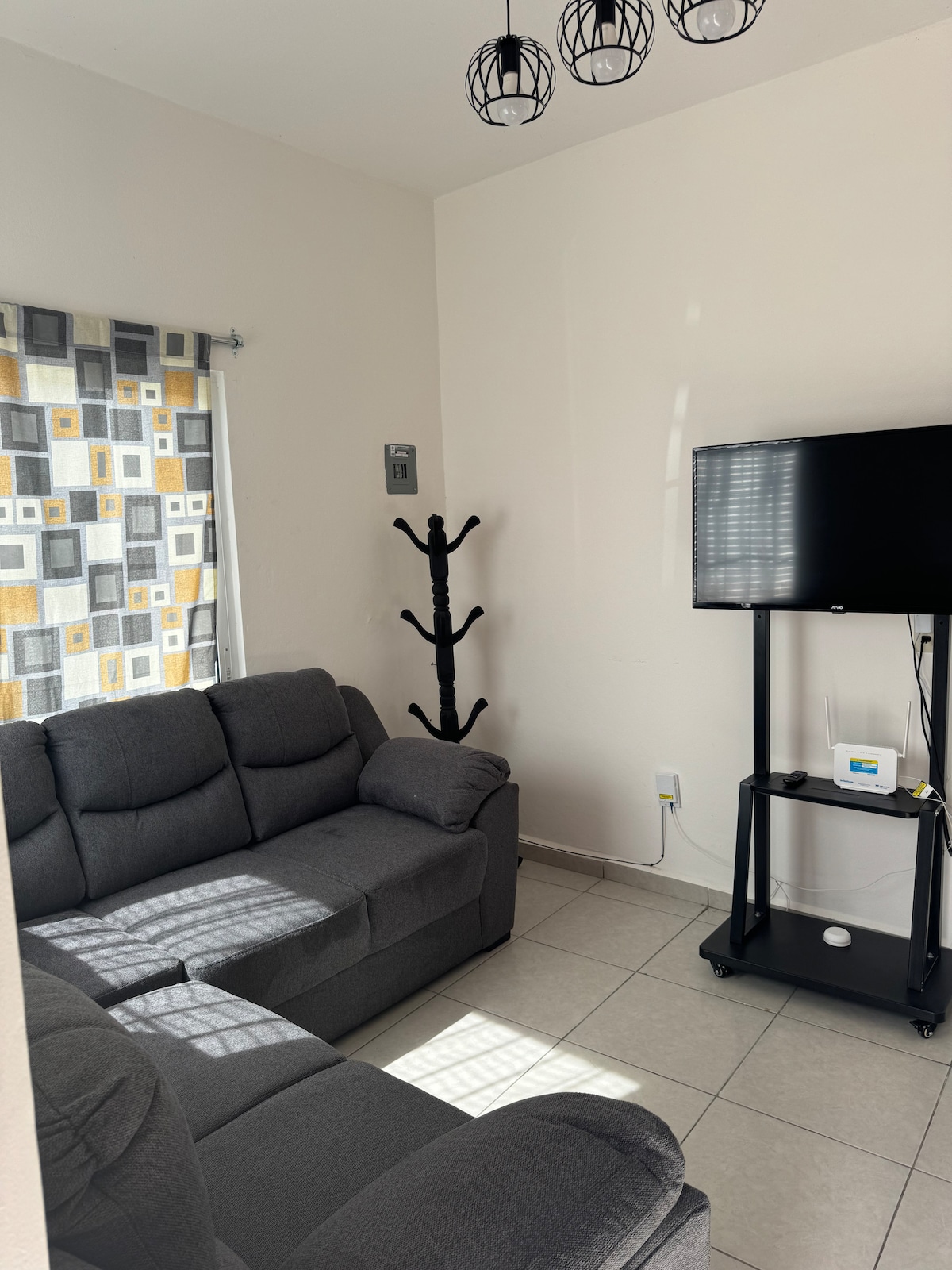 A light-filled living area features a comfortable gray sofa positioned near a mounted television on a mobile stand. A coat rack stands in the corner, and a window is adorned with a patterned curtain, introducing a subtle pop of color.