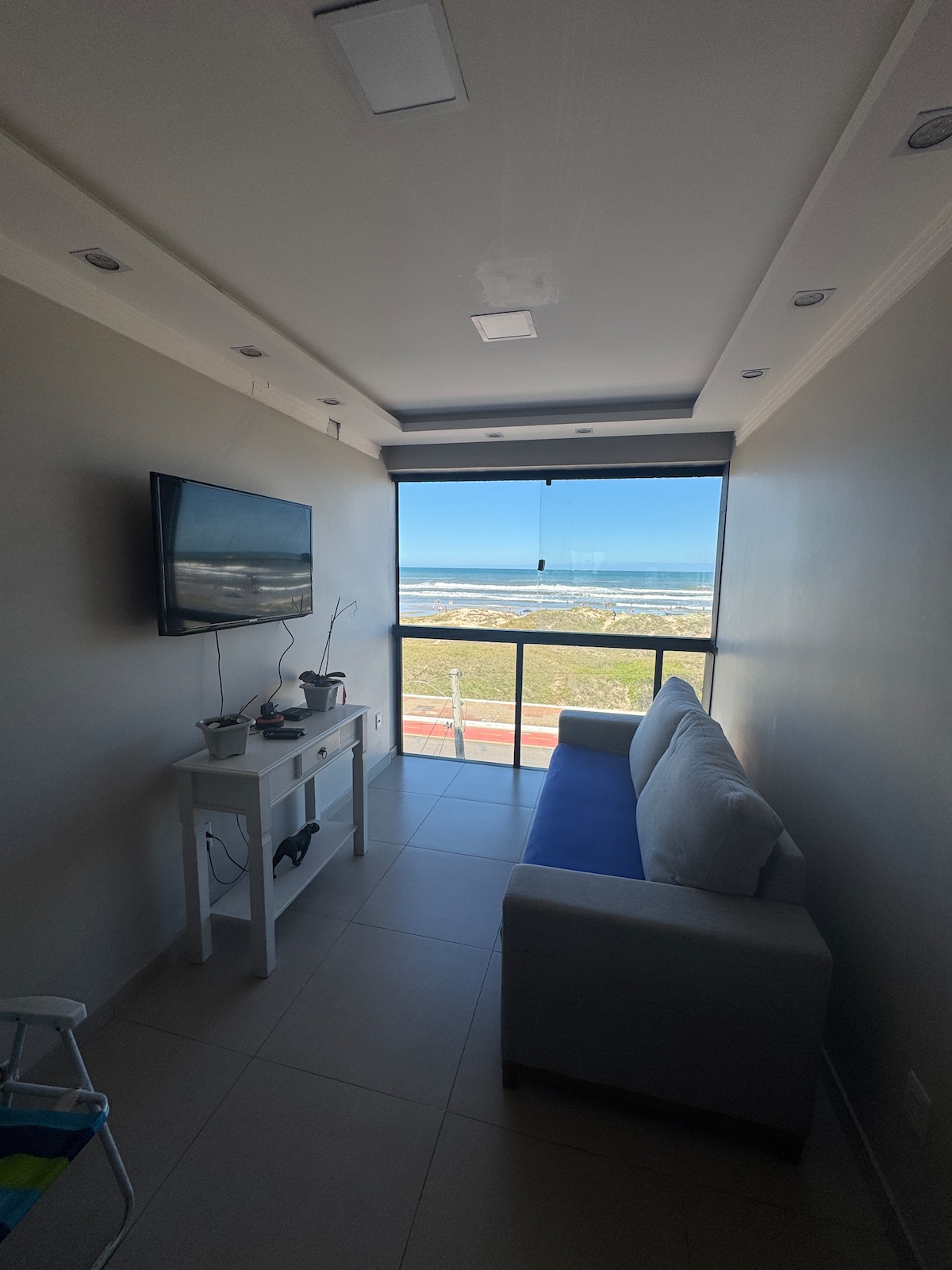 A well-lit space is showcased, featuring a gray sofa positioned along a wall. A flat-screen TV is mounted on the opposite wall, above a narrow console table. Floor-to-ceiling windows provide a clear view of the landscape, allowing natural light to enhance the area.
