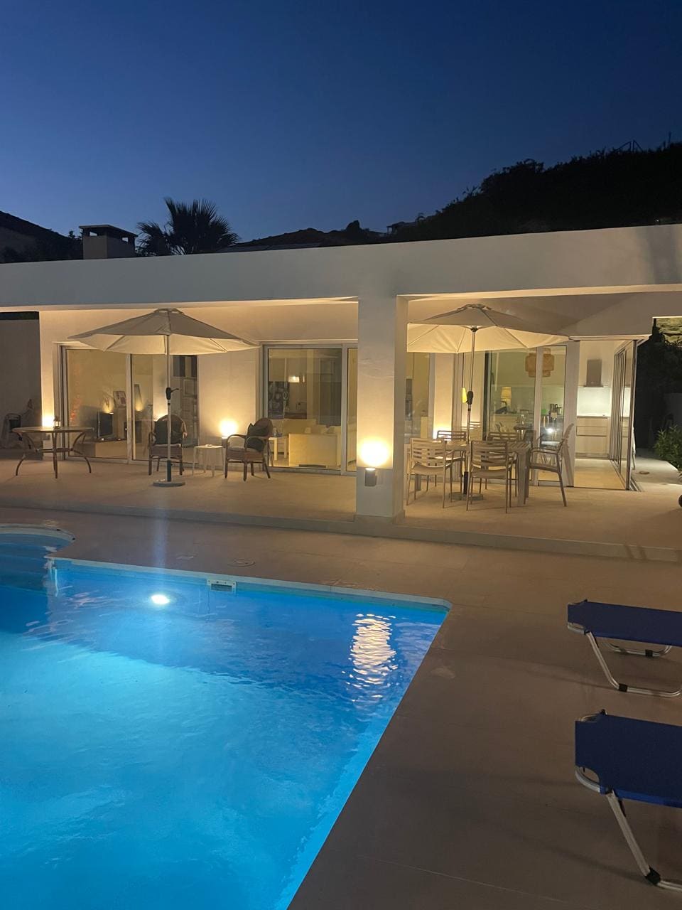 The image captures a modern bungalow illuminated at night, revealing a private swimming pool reflecting soft lights. A spacious terrace features outdoor seating under large umbrellas, while large glass doors provide a view into the dining area, blending indoor and outdoor living.