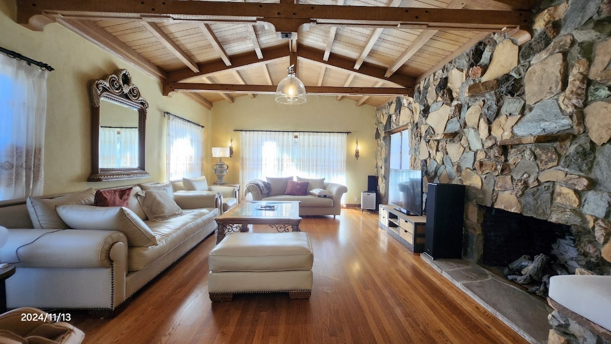 The living area features a stone accent wall and wooden beams above. A light-colored sofa and chairs are arranged around a central coffee table, with a large mirror reflecting natural light. A television is positioned against the opposite wall, enhancing the open space.