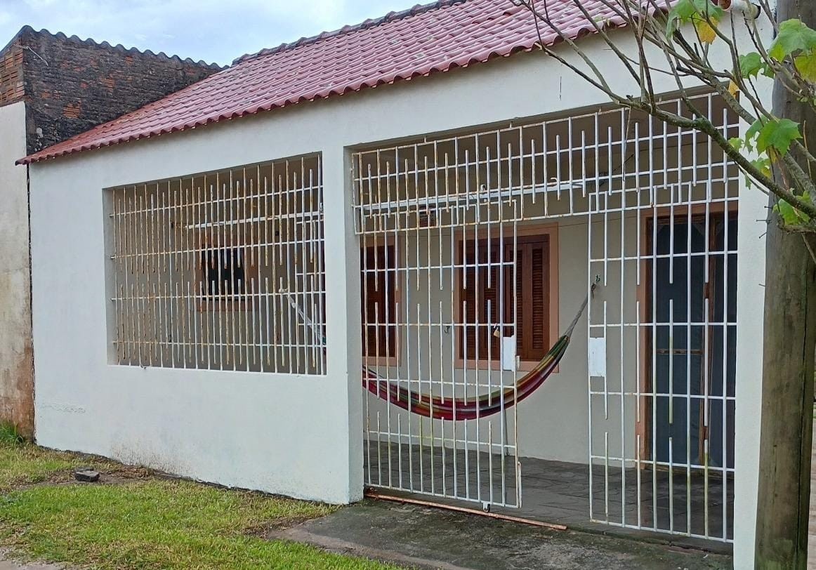 The exterior of a single-story house is shown, featuring a gated entrance with horizontal bars. A colorful hammock is suspended in the front space. Light brown doors are visible, framed by wooden accents, and a grassy area is present, enhancing the sense of outdoor enjoyment.