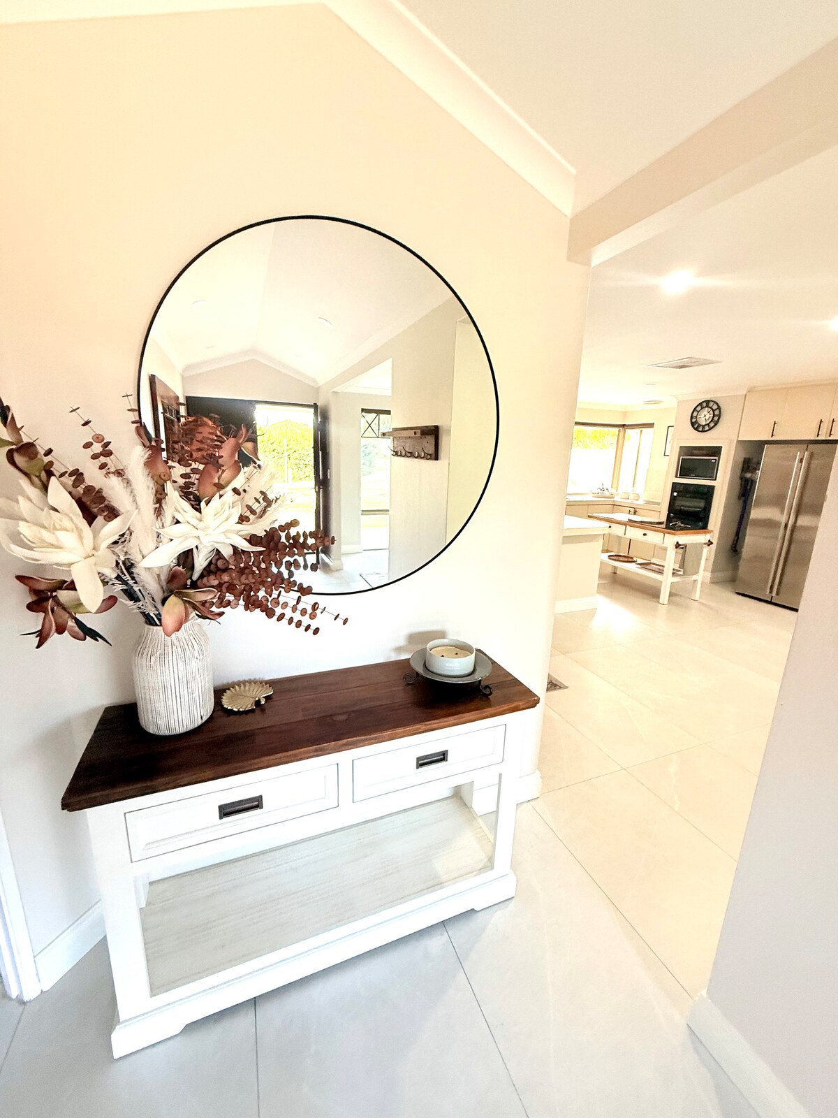 A bright entryway showcases a modern mirror above a wooden console table. A decorative vase with dried foliage adds a touch of nature. Soft lighting illuminates the space, leading to an open kitchen and dining area visible in the background.