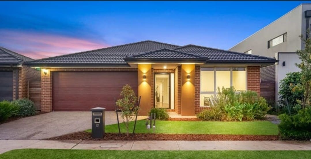 A contemporary family home is displayed, featuring a well-manicured front yard with lush greenery and a welcoming entryway. The warm exterior lighting highlights the brick facade, complemented by a stylish garage door. A tranquil sky serves as a picturesque backdrop.