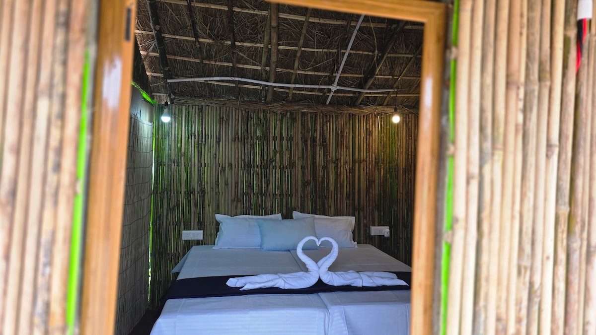 A cozy bamboo cottage is viewed through an open doorway. Inside, a spacious bed with crisp white linens and decorative towels arranged in a heart shape creates a serene atmosphere. Textured bamboo walls and soft lighting contribute to a calming retreat.