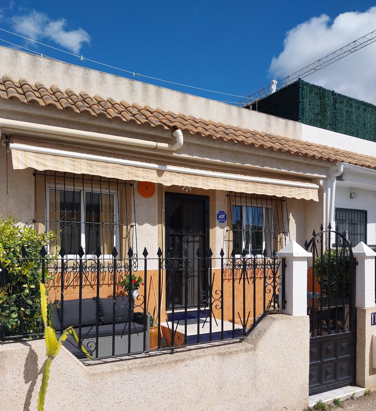 A welcoming façade of a house is framed by a decorative black iron gate and a low wall with vibrant greenery. Above the entrance, a striped awning offers shade. An inviting seating area is positioned on the porch, blending functionality with outdoor charm.