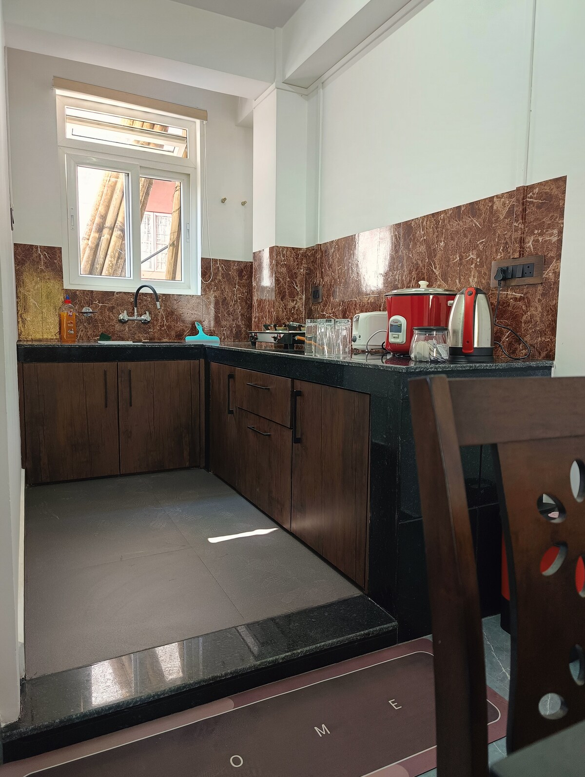 A modern kitchen is visible, featuring dark wood cabinetry and a contrasting black countertop. Natural light enters through a window, illuminating the clean space. Kitchen appliances, including a red kettle and a small dish rack, are neatly arranged. The floor is tiled with a solid black surface.