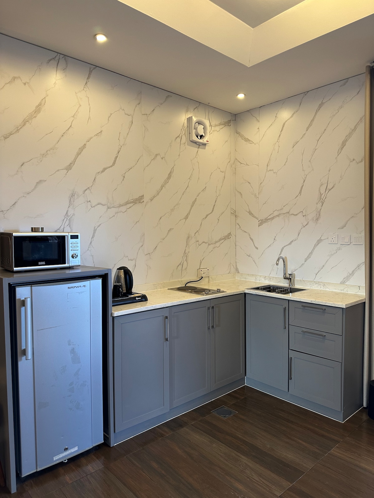 A modern kitchen area is equipped with sleek gray cabinetry and a light marble backsplash. A microwave and kettle are visible on the countertop, alongside a small sink. A refrigerator stands on one side, completing the functional space.