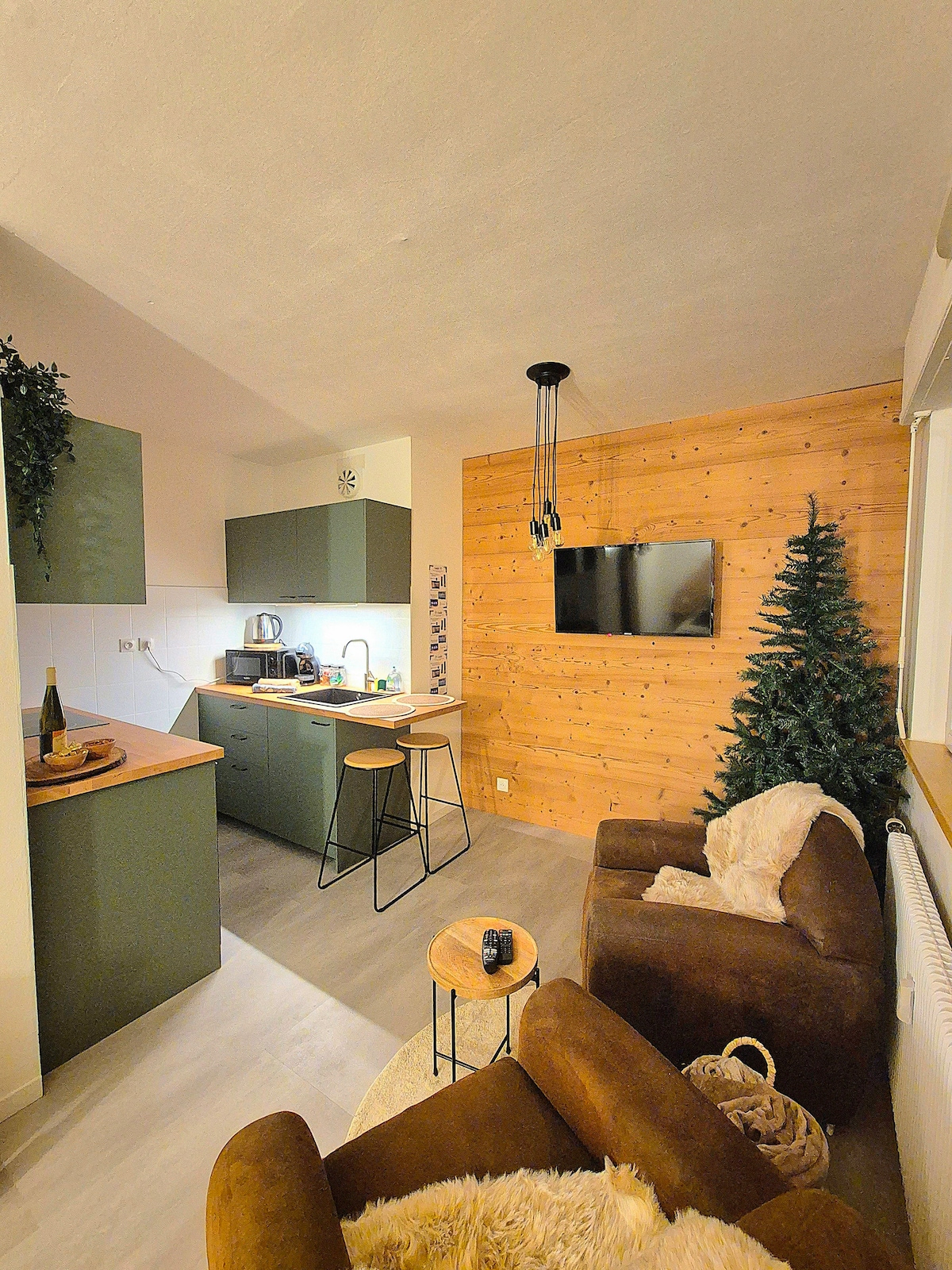 A modern living area features a wooden accent wall and a small Christmas tree. The kitchen showcases minimalist cabinetry and a countertop with bar stools. A comfortable armchair and side table add to the cozy ambiance, while a wall-mounted television offers entertainment options.