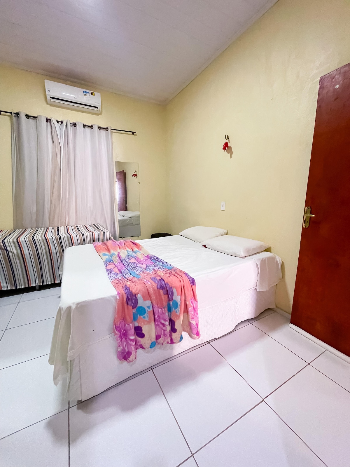 A bright room features a spacious bed adorned with a vibrant blanket. Light-colored walls create an airy atmosphere. A striped bed with decorative pillows is positioned nearby. Natural light enters through a window covered by sheer curtains, while a mirrored wall reflects the cozy interior.