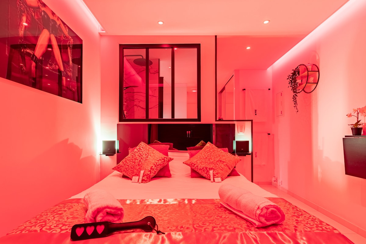 The intimate sleeping area features a spacious bed adorned with decorative pillows and soft towels. Warm red lighting creates a cozy ambiance, complemented by sleek black accents. A stylish mirror and artwork add visual interest, while subtle details enhance the overall decor.