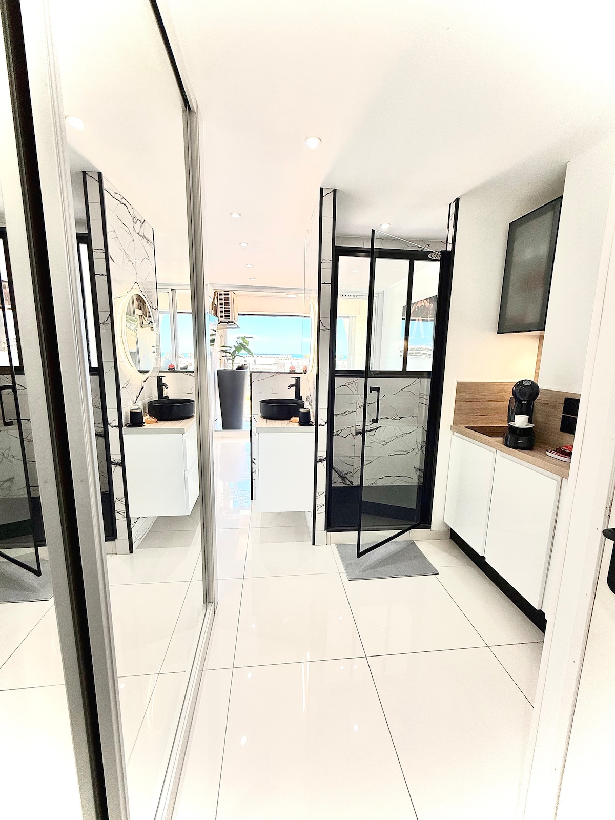 A modern bathroom is shown, featuring a sleek glass shower with a view through its transparent walls. Stylish cabinetry and a mirror reflect the bright, tiled flooring and contemporary design. Natural light enters from a nearby window, enhancing the spacious feel of the area.