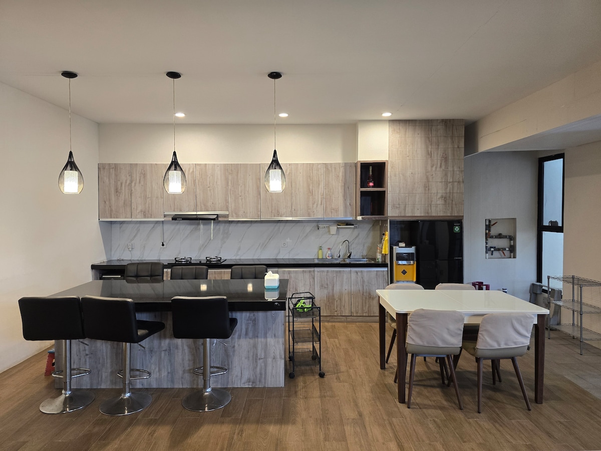 The spacious kitchen features modern cabinetry and a sleek countertop with bar stools. Pendant lights illuminate the area, which includes a dining table surrounded by four chairs. A refrigerator and kitchen appliances are visible, enhancing functionality in the open layout.