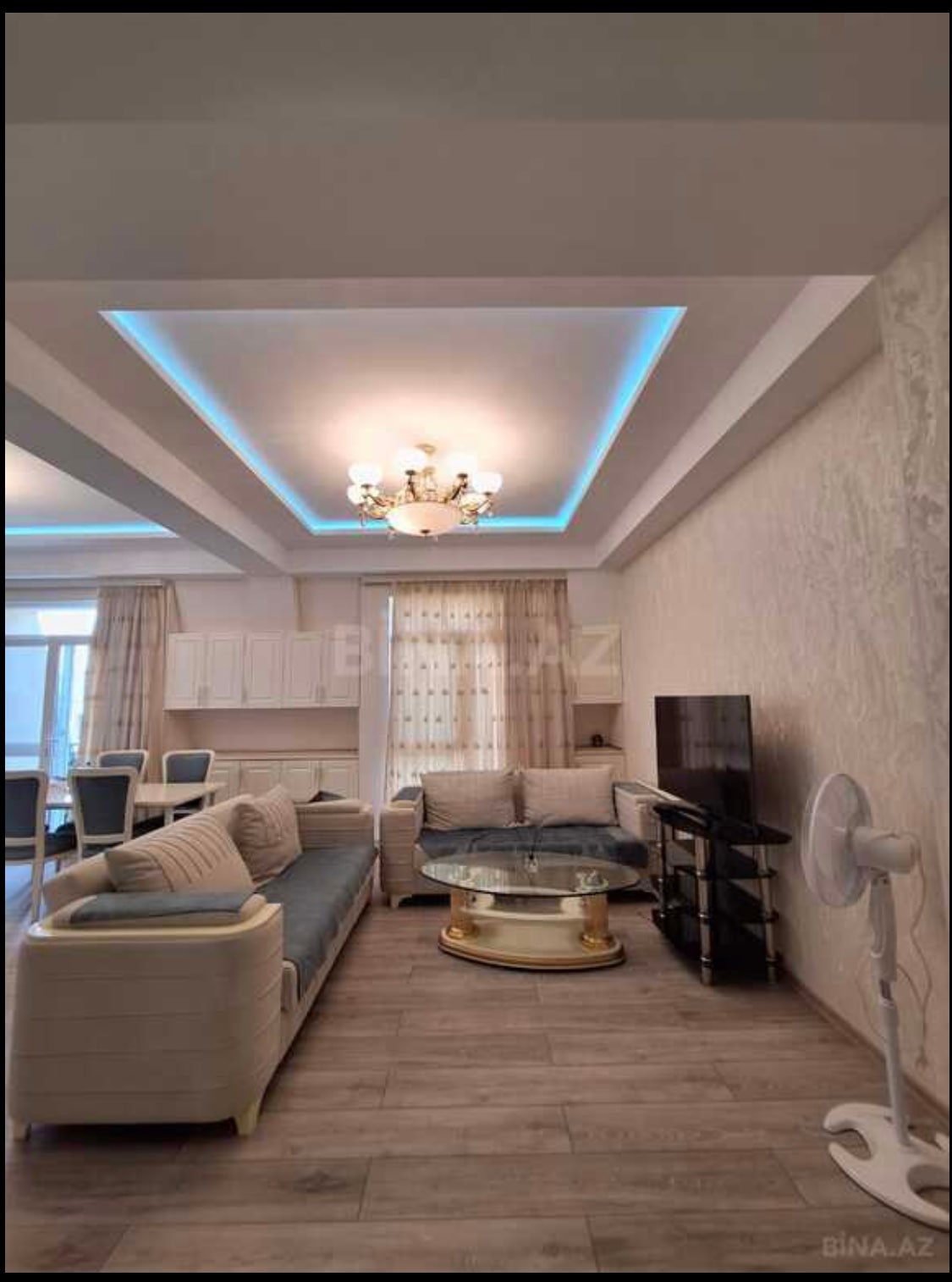 A modern living area is shown, featuring a comfortable sofa set and glass coffee table. A TV is mounted on the wall opposite the seating. Natural light enters through large windows, with light curtains framing the view. Subtle blue accent lighting enhances the ceiling.