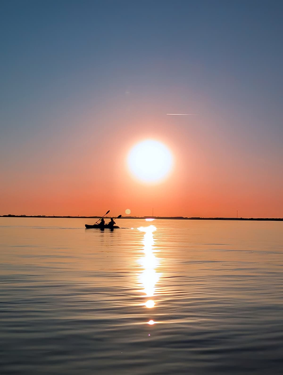 A serene lake scene captures two individuals in a kayak, paddling across smooth water at sunset. The sun casts a warm glow, creating sparkling reflections on the surface, with the horizon lined by distant land silhouettes.