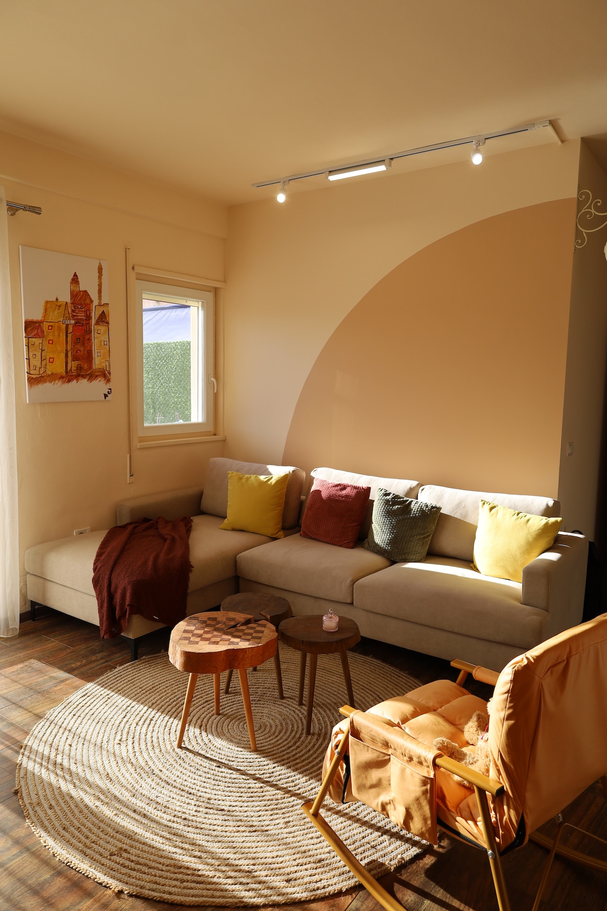 A cozy living area is presented with a sectional sofa adorned with cushions in various hues. A neutral-colored area rug lays beneath a small table. Natural light enters through a window, enhancing the soft tones of the curved wall art.