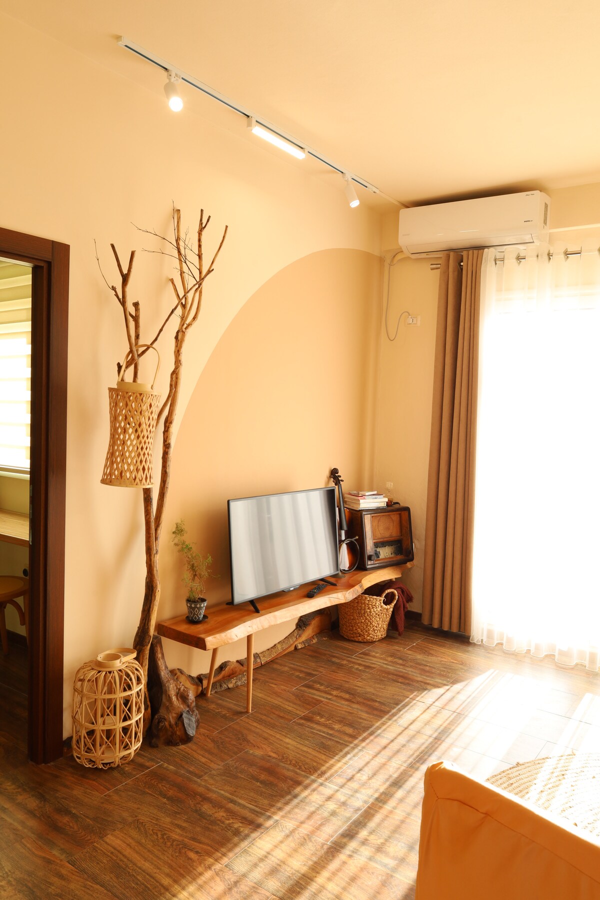 A cozy living area features a television mounted on a wooden stand. Natural light floods in through a large window with sheer curtains. Decorative elements include a wicker hanging planter and a tree branch accent. The warm tones of the walls create a welcoming ambiance.