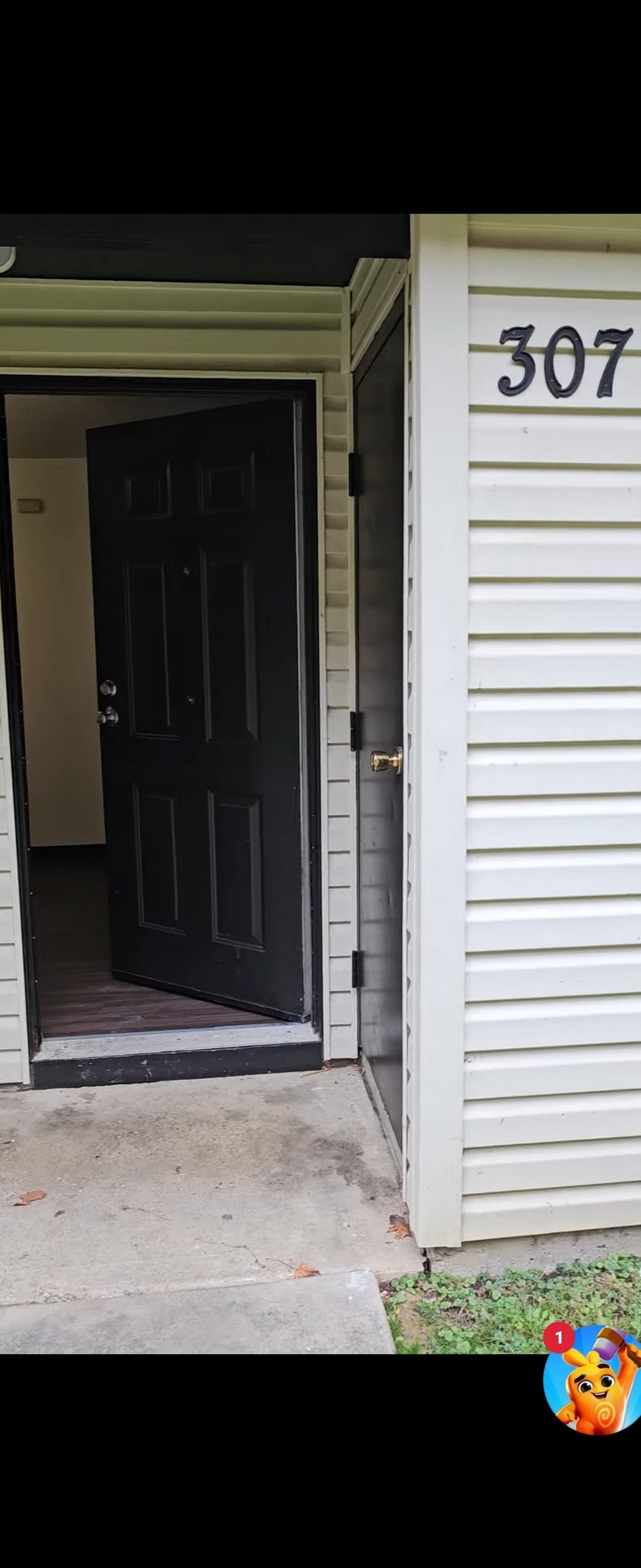 The entrance to the townhouse is shown with an open black front door, revealing an interior space. The door is framed by a light-colored exterior wall, and the unit number '307' is displayed nearby. A small concrete path leads up to the entrance.