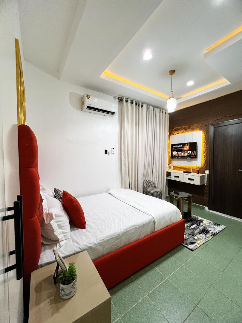 The studio features a spacious bed with a red headboard and crisp white bedding. A small table sits beside the bed, and light-colored curtains frame a window. A mounted TV is visible on the wall opposite, with soft lighting highlighting the minimalist décor.