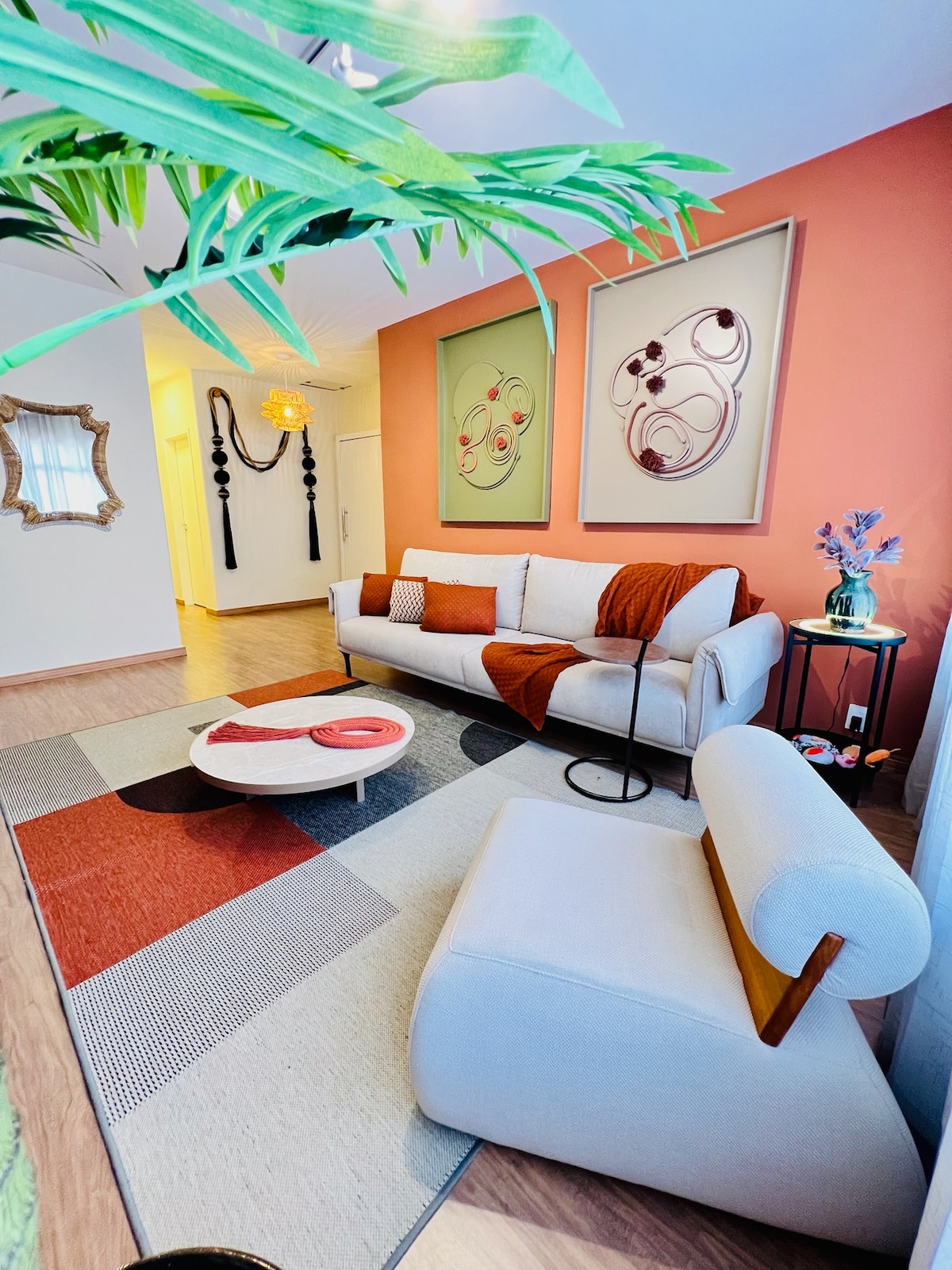 The living area features a light-colored sofa with a burgundy throw blanket and a small side table. A colorful area rug with geometric patterns extends across the floor. Framed artwork adorns the walls, and a plant adds a touch of greenery to the space.