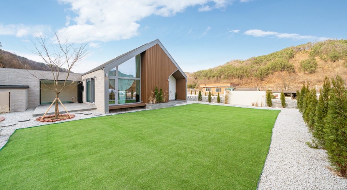 A spacious yard features lush artificial grass, providing ample area for leisure. The modern single-story structure showcases a distinctive angled roof, with large windows allowing natural light to enter. Nearby, a decorative gravel path complements the well-maintained landscaping.