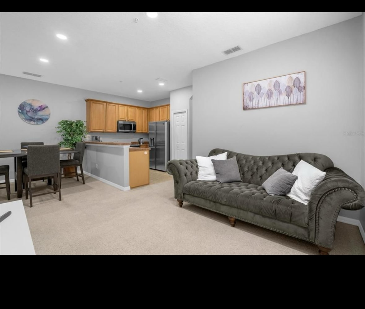 A comfortable living area features a tufted sofa adorned with pillows. A dining table is positioned nearby, complemented by wooden cabinetry. The kitchen area is visible in the background, equipped with modern appliances. Neutral tones and soft carpet create a homely environment.