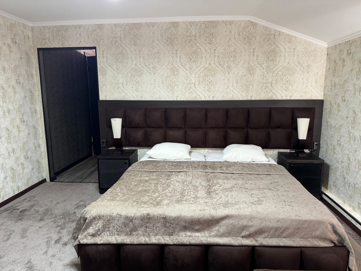 A spacious sleeping area is presented with a large tufted headboard above a neatly arranged bed. Soft lighting is provided by two bedside lamps on dark nightstands. The textured wallpaper and carpet create a cohesive design, contributing to a comfortable atmosphere.